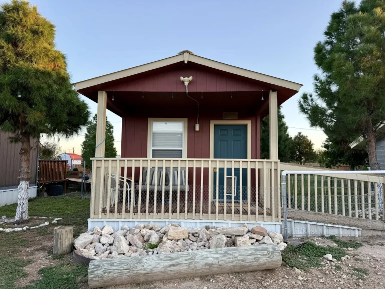 A small, dog-friendly red cabin in Garden City, TX, with a covered front porch, a dark green door, and a wooden ramp.