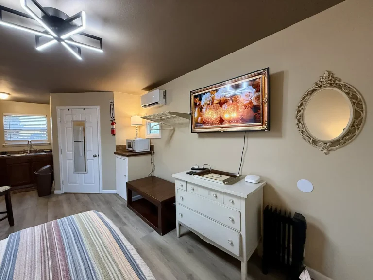 A white dresser, an ornate oval mirror, and a wall-mounted TV displaying glowing pumpkins in a dog friendly cabin in Garden City, TX, with a glimpse of a kitchen and a striped bed in the foreground.