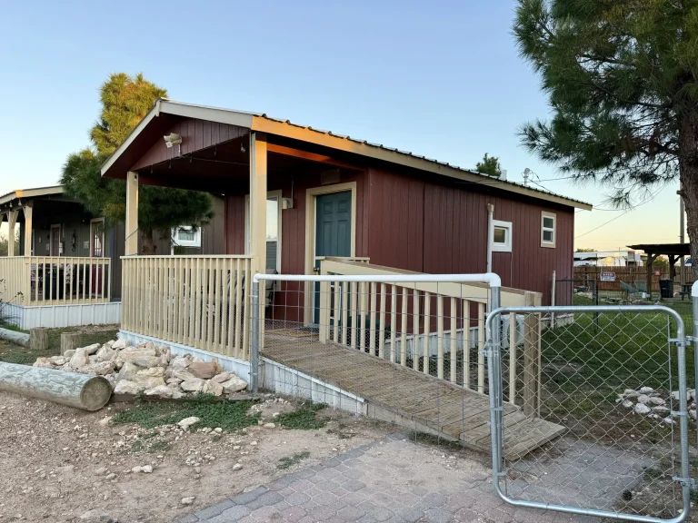 A maroon, dog-friendly cabin in Garden City, TX, featuring a covered front porch and a wooden accessibility ramp leading to a green door.
