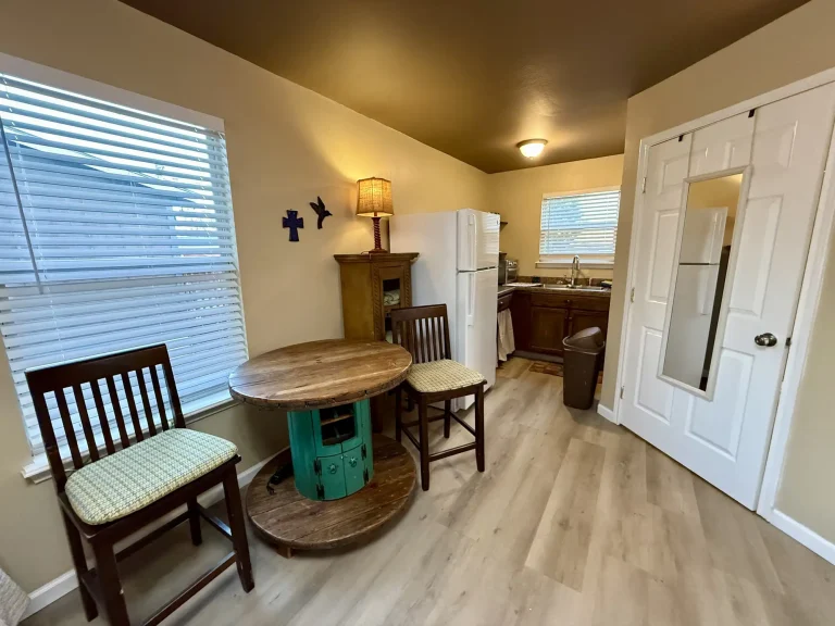 A small kitchen and dining area in a dog friendly cabin featuring a round wooden table made from a repurposed cable spool, two wooden chairs with patterned cushions, and a white refrigerator.