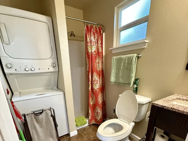 A bathroom in a dog friendly cabin featuring a stacked washer and dryer, a shower with a patterned red curtain, a toilet, and a vanity sink.